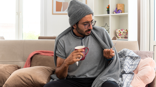 Man sitting on a couch holding a mug, wrapped in a blanket in a cozy living room.